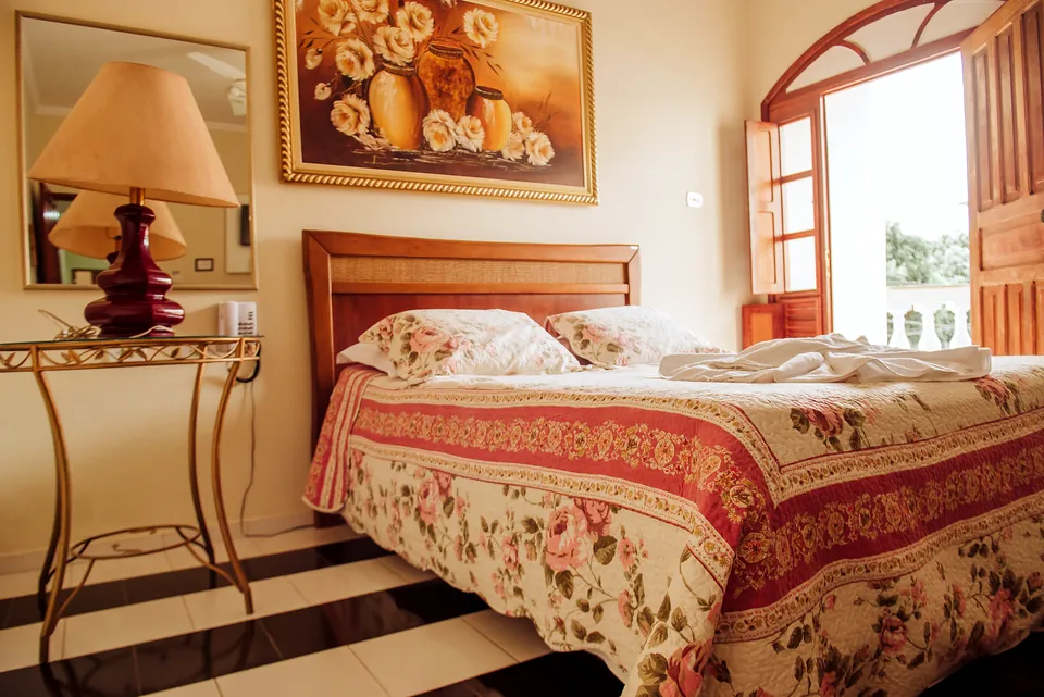Quarto Casal com cama de casal de madeira, colcha florida, abajur vermelho e janela em arco
