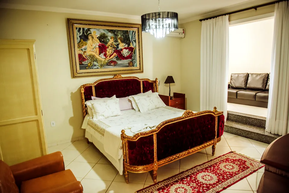Suíte Master com lustre de cristal, cama de cabeceira vermelha e quadro de moldura dourada